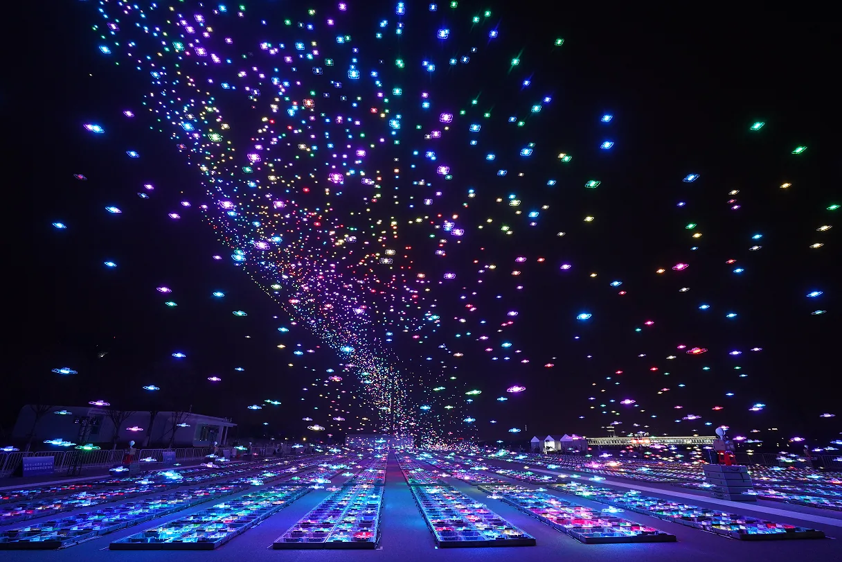 Cientos de drones despegando en formación con luces multicolor, creando una espiral de luz en el cielo nocturno